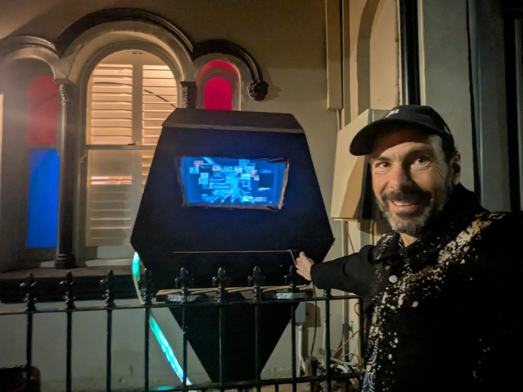 The Algorithm — a black, coffin-shaped cabinet with a glowing screen and LED strip, installed on a porch at night.