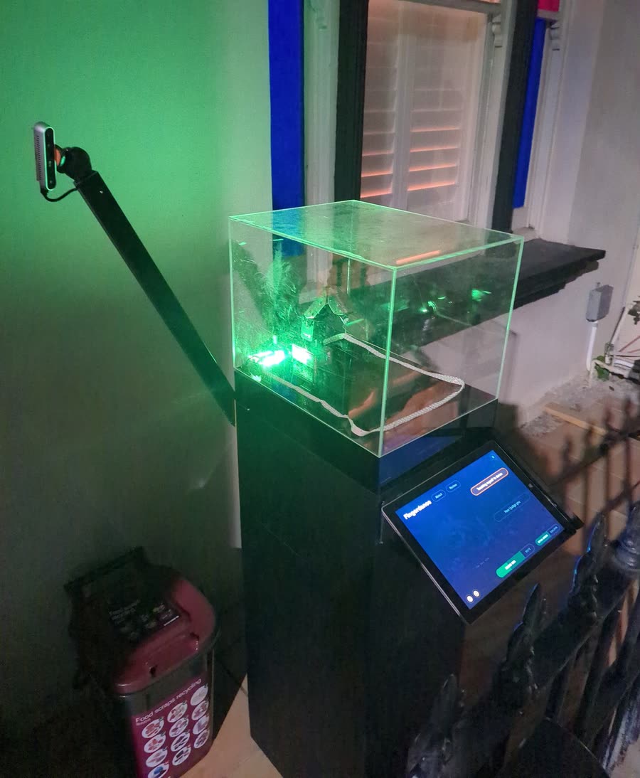 Fingerdance on the porch at night: a robot finger in a clear case on a black plinth, lit green, with a depth camera on a pole beside it and a tablet angled on the front.